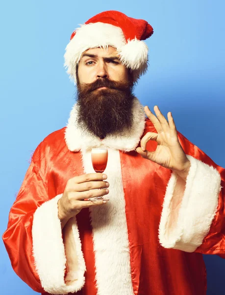 ハンサムbeadedサンタクラス男ともに長いです髭で面白いです顔ホールディングガラスのアルコールショットで赤クリスマスやクリスマスセーターと新しい年の帽子ショーok上の青スタジオ背景 — ストック写真