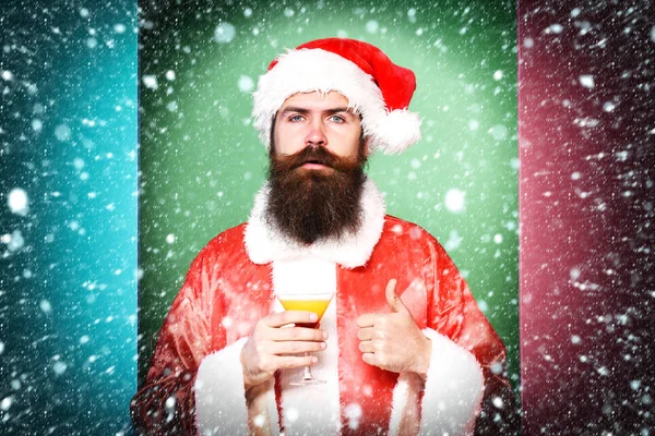 잘생긴 크리스마스 또는 크리스마스 스웨터와 멋진 다채로운 스튜디오 배경에서 보여주는 새 해 모자에 알콜 음료 잔을 들고 심각한 얼굴에 긴 수염을 가진 산타 클로스 남자 수염 — 스톡 사진