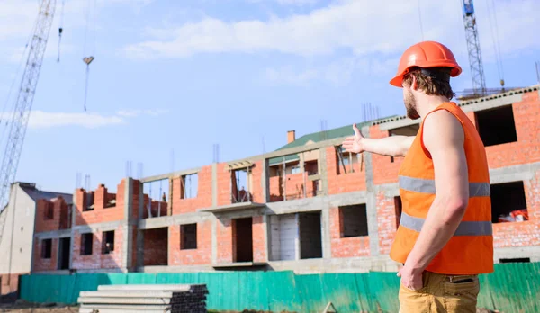 Chaleco constructor y sitio de construcción del casco apuntando al edificio. Concepto de responsabilidades del contratista. Casco de protección Guy de pie en frente del edificio hecho de ladrillos. Proceso de construcción control — Foto de Stock