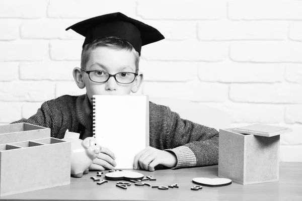 Νεαρός μαθητής χαριτωμένος αγόρι στο γκρι πουλόβερ και τα γυαλιά, κάθεται στο γραφείο με copybook στο χέρι ξύλινοι αριθμοί ροζ χοίρου γουρουνάκι Τράπεζα στο καπάκι αποφοίτηση τάξη σε φόντο λευκό τούβλο τοίχο, αντίγραφο spac — Φωτογραφία Αρχείου