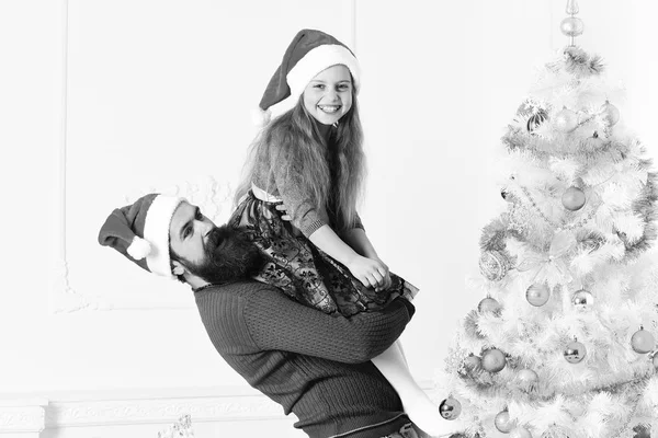 Christmas eve and New Year concept. Girl and bearded man with happy faces near blue and white Christmas tree on white background. Father holds little daughter on arms. Kid and Santa decorate fir tree