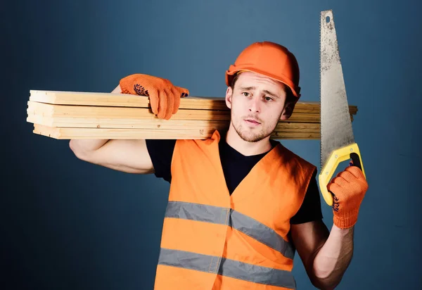 Menuisier, menuisier, ouvrier, constructeur sur un visage réfléchi porte des poutres en bois sur l'épaule. Concept de Woodcraft. Homme, homme à tout faire avec casque, scie à main et poutres en bois, fond gris — Photo