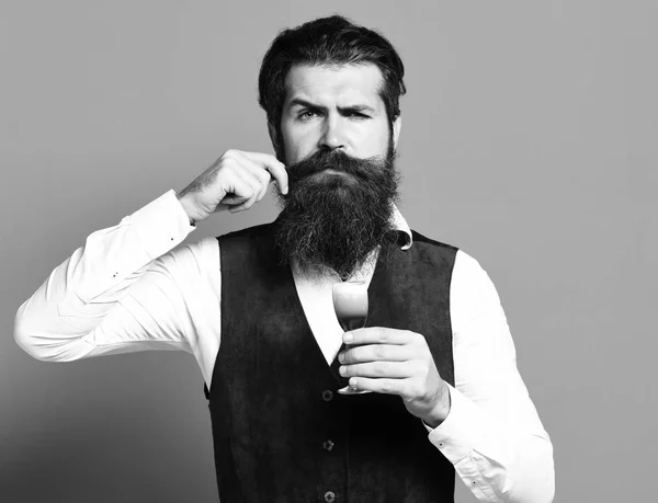 Bel homme barbu avec longue barbe et moustache sur le visage sérieux tenant verre de coup alcoolique en daim vintage gilet en cuir sur fond rouge studio — Photo