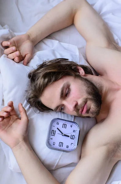 Man bearded unshaven handsome guy lay on pillow near alarm clock top view. Guy relaxing bed before alarm clock ringing. Toughest part of morning simply getting out of bed. Get up early morning tips