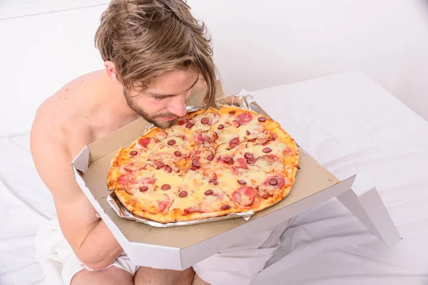 L'homme aime la pizza pour le petit déjeuner. Nutrition pour célibataires. Homme barbu beau gars manger de la nourriture au fromage pour le petit déjeuner au lit. Le gars tient la boîte à pizza assis lit dans la chambre ou la chambre d'hôtel. Service de livraison de nourriture — Photo