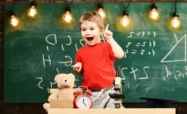 Kid boy near microscope in classroom, chalkboard on background. First former excited about studying, learning, education. Enthusiastic pupil concept. Child with inspired expression near microscope — Stock Photo, Image