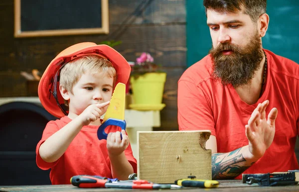 Niño, niño ocupado en casco protector aprendiendo a usar la sierra manual con papá. Concepto de juegos educativos. Padre, padre con barba enseñando a su hijo a serrar mientras su hijo juega con la sierra de juguete — Foto de Stock