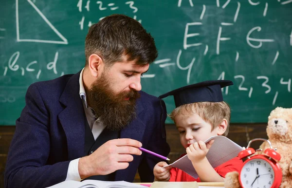 Father with beard, teacher teaches son, little boy. Individual schooling concept. Kid studies individually with teacher, at home. Teacher and pupil in mortarboard, chalkboard on background