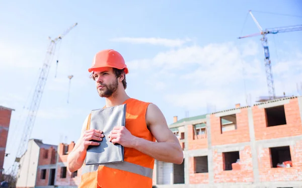 Control del contratista según el plan. Casco de protección Guy pie en frente del edificio hecho de ladrillos rojos. Proceso de construcción de control. Constructor naranja chaleco y obras de casco en el sitio de construcción — Foto de Stock