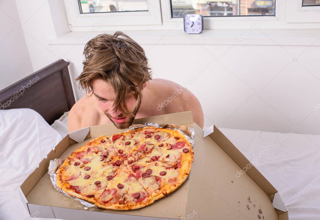 Who cares about diet. Man bearded handsome guy eating food pizza for breakfast in bed. Guy holds pizza box sit bed in bedroom or hotel room. Food delivery service. Man likes pizza for breakfast