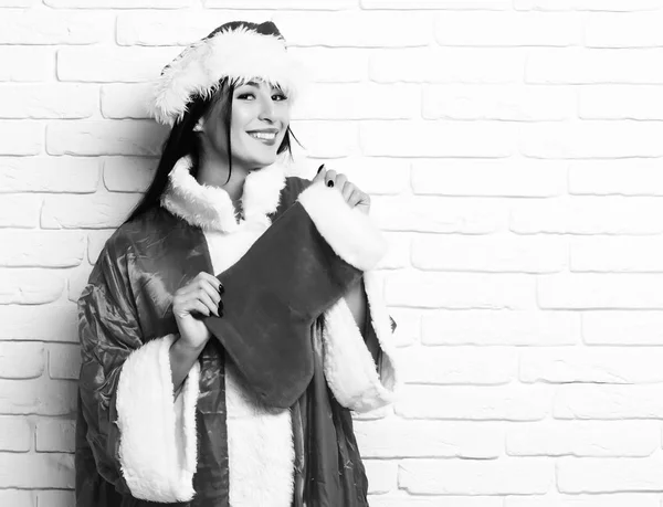 Vrij leuk sexy santa meisje of lachende brunette vrouw in trui Nieuwjaar en Kerstmis of xmas hoed houdt decoratieve kous of boot op witte bakstenen muur achtergrond — Stockfoto