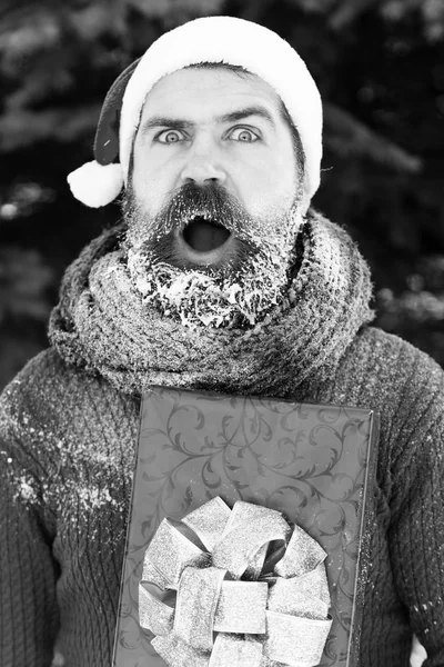 Handsome surprised man in santa claus hat, bearded hipster with beard and moustache covered with white frost, holds red gift box on sunny winter day outdoors on natural backgroun — Stock Photo, Image