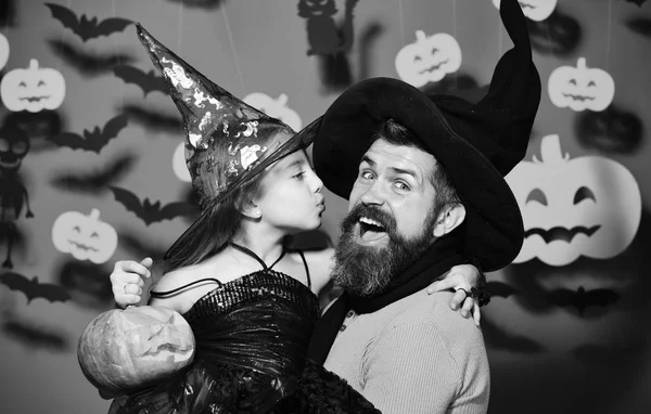Wizard and little witch in black hats hold pumpkin. Halloween party and family concept. Girl kisses bearded man with happy face on red background with decor. Father and daughter in Halloween costumes