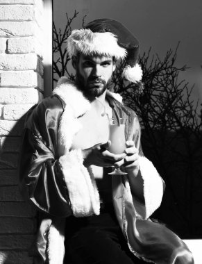 Genç, yakışıklı, sakallı, seksi Noel Baba sakallı, kırmızı Noel Baba şapkalı ve ceketli, kaslı, çıplak gövdeli, siyah ve tuğla duvar arkasında alkollü turuncu bir içecek bardağı tutan adam.