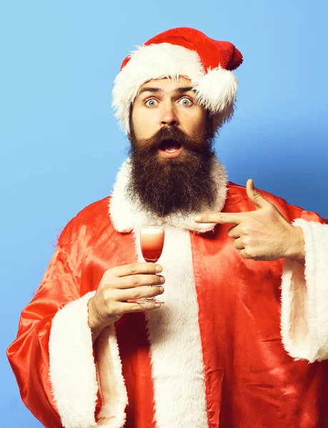 Guapo barbudo hombre de Santa Claus con barba larga en sorprendidocara celebración de vidrio de tiro alcohólico en rojo navidad o suéter de Navidad y sombrero de año nuevo en fondo estudio azul —  Fotos de Stock