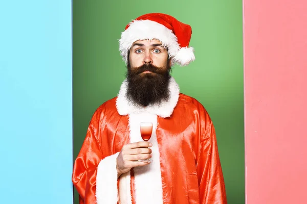 Knappe man van de baard Kerstman met lange baard op verbaasd gezicht houden glas alcoholische schot in rode kerst of xmas vacht en Nieuwjaar hoed op kleurrijke studio achtergrond — Stockfoto