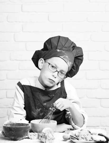 Joven chico lindo cocinero en uniforme rojo y sombrero en la cara manchada con gafas sentadas en la mesa con cuencos de colores, sabrosos pasteles, rodillo y batidor de cocina en fondo de pared de ladrillo blanco — Foto de Stock