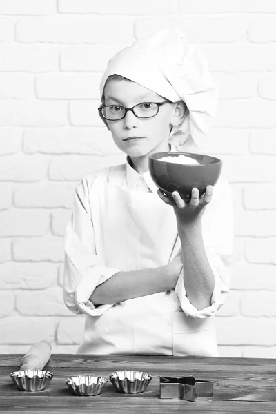 Jeune garçon petit chef cuisinier mignon en uniforme blanc et chapeau sur le visage sérieux avec des lunettes debout près de la table avec des moules à rouleau pour gâteaux et tenant bol rouge sur fond de mur de brique — Photo