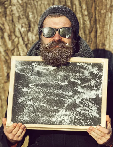 Stilig skäggig man, hipster, med skägg och mustasch i svarta solglasögon täckt med vit frost innehar svarta tavlan eller svarta tavlan vinterdag på naturlig bakgrund, kopia spac — Stockfoto