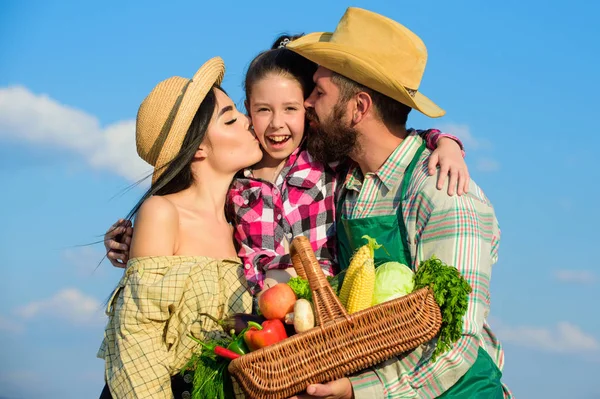 Parents and daughter farmers celebrate harvest holiday. Family farmers hug kiss kid hold basket fall harvest. Family gardener basket harvest blue sky background. Family gardening. Family farm concept