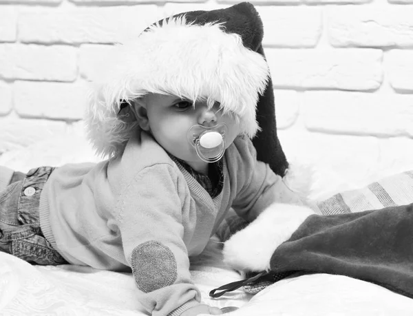Mały chłopiec z uroczą ciekawą twarzą w żółtym swetrze leżącym na brzuchu z manekinem w kapeluszu noworocznym i Boże Narodzenie lub świąteczne pończochy lub buty na białym tle ściany z cegły — Zdjęcie stockowe