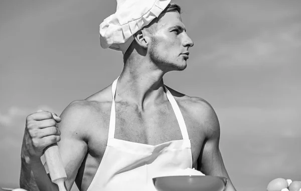 Homme sur le visage confiant porte chapeau de cuisine et tablier, ciel sur fond. Concept de cuisine. Chef cuisinier préparer la pâte pour la cuisson. Cuisinier ou chef avec des épaules musclées sexy et la poitrine tient rouleau à pâtisserie — Photo