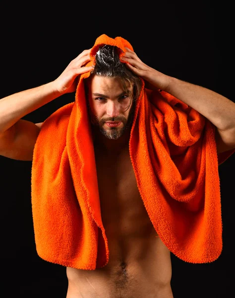 Man bearded tousled hair covered with foam or soap suds. Wash off foam with water carefully. Man with orange towel ready to take shower. Water is over. Macho attractive nude guy black background