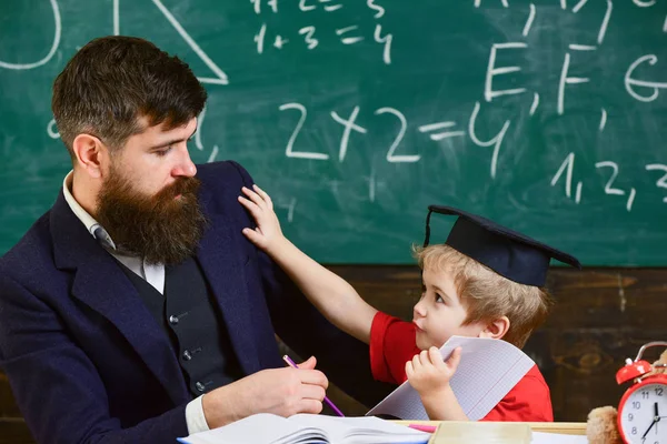 Father with beard, teacher teaches bored son, little boy. Boring studying concept. Kid fed up with studying, kicks away teacher. Teacher and pupil in mortarboard, chalkboard on background
