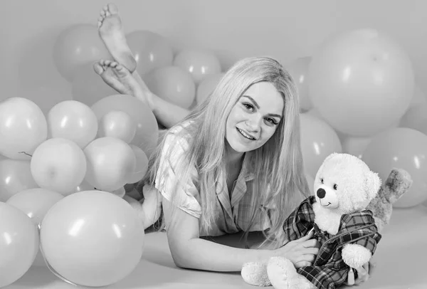 Blonde on smiling face relaxing with teddy bear toy. Girl in pajama, domestic clothes lay near air balloons, pink background. Birthday girl concept. Woman cute celebrate birthday with balloons.