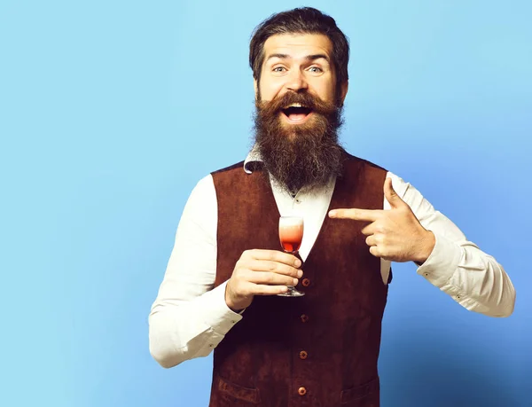 handsome bearded man with long beard and mustache on smiling face tasting glass of alcoholic shot in vintage suede leather waistcoat on blue studio background,copy spac