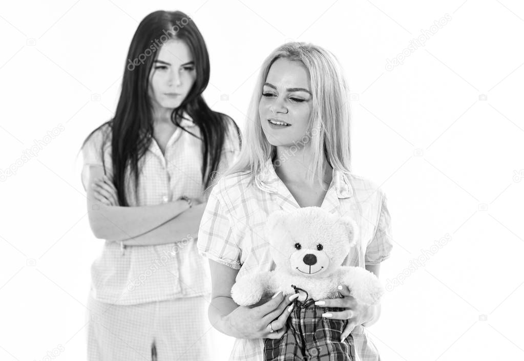 Jealousy and envy concept. Sisters in pajamas looks unfriendly, jealous. Girls in pink pajamas, isolated white background. Lady jealous to sister with plush toy bear, defocused