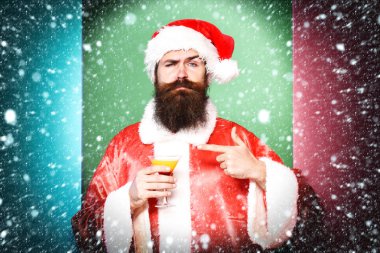 ciddi yüz alkollü içecek bardak Noel veya xmas kazak ve yeni yıl şapka tutan uzun sakallı yakışıklı sakallı Noel Baba adam üzerinde renkli stüdyo arka plan üzerinde gösterir