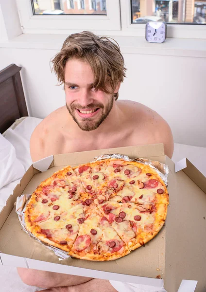 Food delivery service. Man bearded handsome bachelor eating cheesy food for breakfast in bed. Man likes pizza for breakfast. Break diet concept. Guy holds pizza box sit bed in bedroom or hotel room