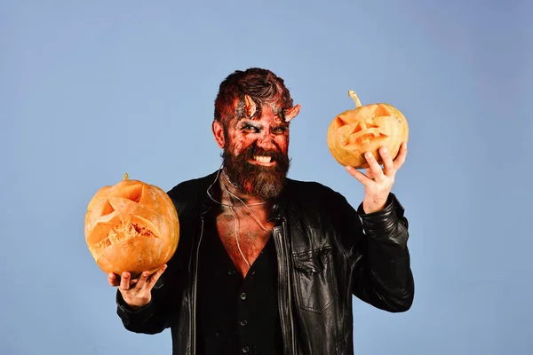 Man wearing scary makeup holds pumpkins on blue background.