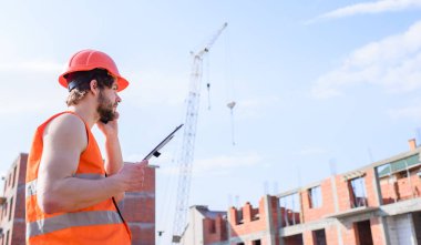 Adam koruyucu kask standı önünde kırmızı tuğla dışında yapılan binanın. Kontrol inşaat süreci. Oluşturucu Turuncu yelek ve kask inşaat alanında çalışır. Yüklenici denetim planına göre