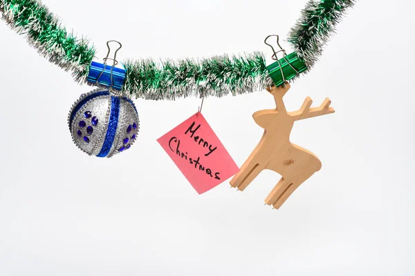 Feliz concepto de Navidad. Hojalata con adornos de Navidad fijados y papel con nota Feliz Navidad, fondo blanco. Bola con adornos y juguete de ciervo de madera cuelgan de oropel verde brillante —  Fotos de Stock