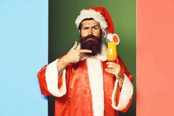 Knappe man van de baard Kerstman met lange baard op verbaasd gezicht houden glas cocktail in rode kerst of xmas trui en Nieuwjaar hoed op kleurrijke studio achtergrond — Stockfoto