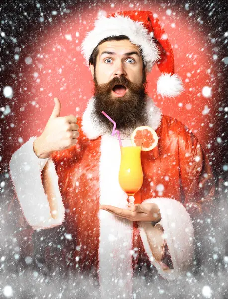 Guapo barbudo hombre de Santa Claus con barba larga en la cara sorprendida sosteniendo vaso de cóctel sin alcohol en navidad o suéter de Navidad y sombrero de año nuevo que muestra fresco en el fondo del estudio rojo —  Fotos de Stock