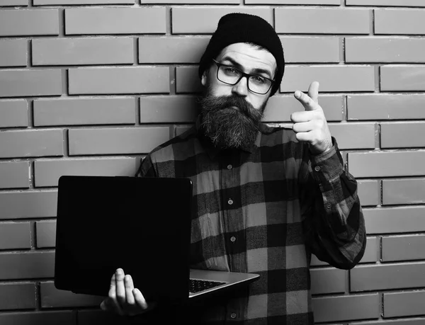 Bearded man, long beard. Brutal caucasian serious unshaven hipster holding laptop in red black checkered shirt with hat and glasses on brown brick wall studio backgroun