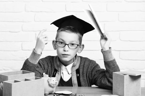Unga söta elev pojke i grå tröja och glasögon som sitter vid skrivbord med häfte i hand trä siffror rosa grisen gris bank i examen keps i klassrummet på vitt tegel vägg bakgrund — Stockfoto