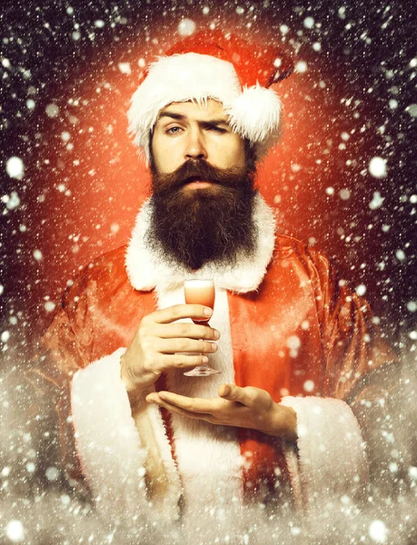 Guapo barbudo hombre de Santa Claus con barba larga en la cara seria sosteniendo vaso de trago alcohólico en la mano en navidad o suéter de Navidad y sombrero de año nuevo en fondo rojo estudio —  Fotos de Stock