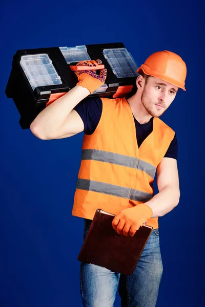 Homem de capacete, chapéu duro segura caixa de ferramentas e pasta com documentos, fundo azul. Conceito de serviço de reparação. Trabalhador, reparador, reparador, construtor em face calma carrega caixa de ferramentas no ombro, pronto para o trabalho — Fotografia de Stock