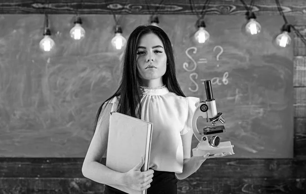 Professor de biologia detém livro e microscópio. Senhora em desgaste formal no rosto calmo na sala de aula. Lady cientista detém livro e microscópio, quadro-negro em segundo plano, espaço de cópia. Conceito de biologia — Fotografia de Stock