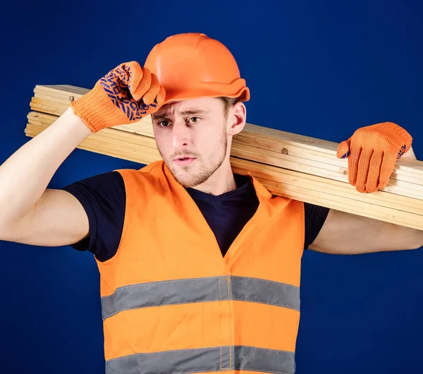 L'homme en gants de protection tient la visière du casque, corrige le casque dur sur la tête, fond bleu. Concept d'équipement de protection. Menuisier, menuisier, ouvrier, constructeur porte une poutre en bois sur l'épaule — Photo