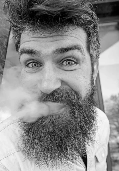 Vaping concept. Hipster met verwarde haren en grijs op ontspannen vrolijk gezicht met witte rook uit mond vliegen. Man met baard en snor roken, gebouw achtergrond — Stockfoto