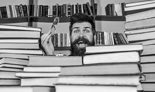 Hombre en la cara feliz entre montones de libros en la biblioteca, estanterías en el fondo. Concepto de investigación científica. Profesor o estudiante con barba usa anteojos, se sienta a la mesa con libros, desenfocado —  Fotos de Stock