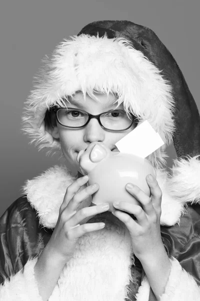 Joven lindo santa claus chico con gafas en suéter rojo y año nuevo sombrero de Navidad celebración y besos rosa alcancía sobre fondo de estudio azul —  Fotos de Stock