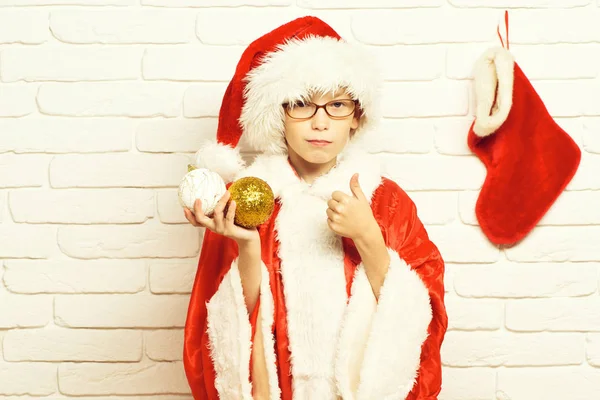 若いかわいいサンタ クロース メガネ少年は赤いセーターを着て、新年帽子を示すクールなと保持している装飾的なクリスマスやクリスマスのストッキングと白いレンガ壁の背景にブートのカラフルなボールで — ストック写真