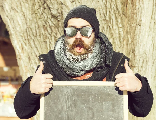 Homem animado, hipster, com barba e bigode em óculos escuros cobertos com geada branca dá polegares duplos até gesto com placa preta no dia de inverno no fundo natural, cópia spac — Fotografia de Stock