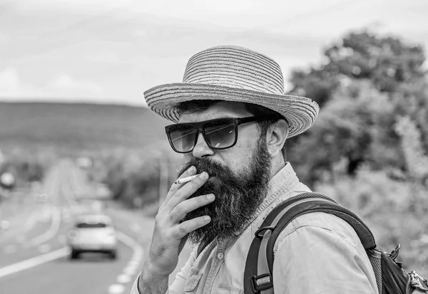 Cigarette before long journey. Traveler stylish hipster take brake.Man with beard and mustache in straw hat smoking cigarette, road background defocused. Smoking cigarette before long journey